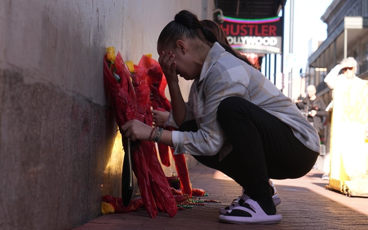 A woman crouches beside a wall, holding a yellow rose wrapped in red tissue paper in one hand, her other hand covering her face. There are other similarly wrapped yellow roses leaning against the wall.