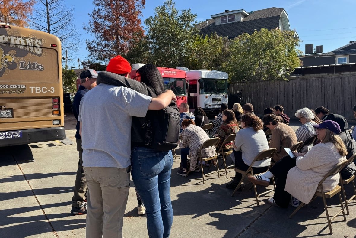 Two people are seen from the back as they stand, embracing each other, next to several rows of chairs filled with people. The back of a yellow bus is visible at the left side of the image, and there are more buses at the right side beyond the chairs.